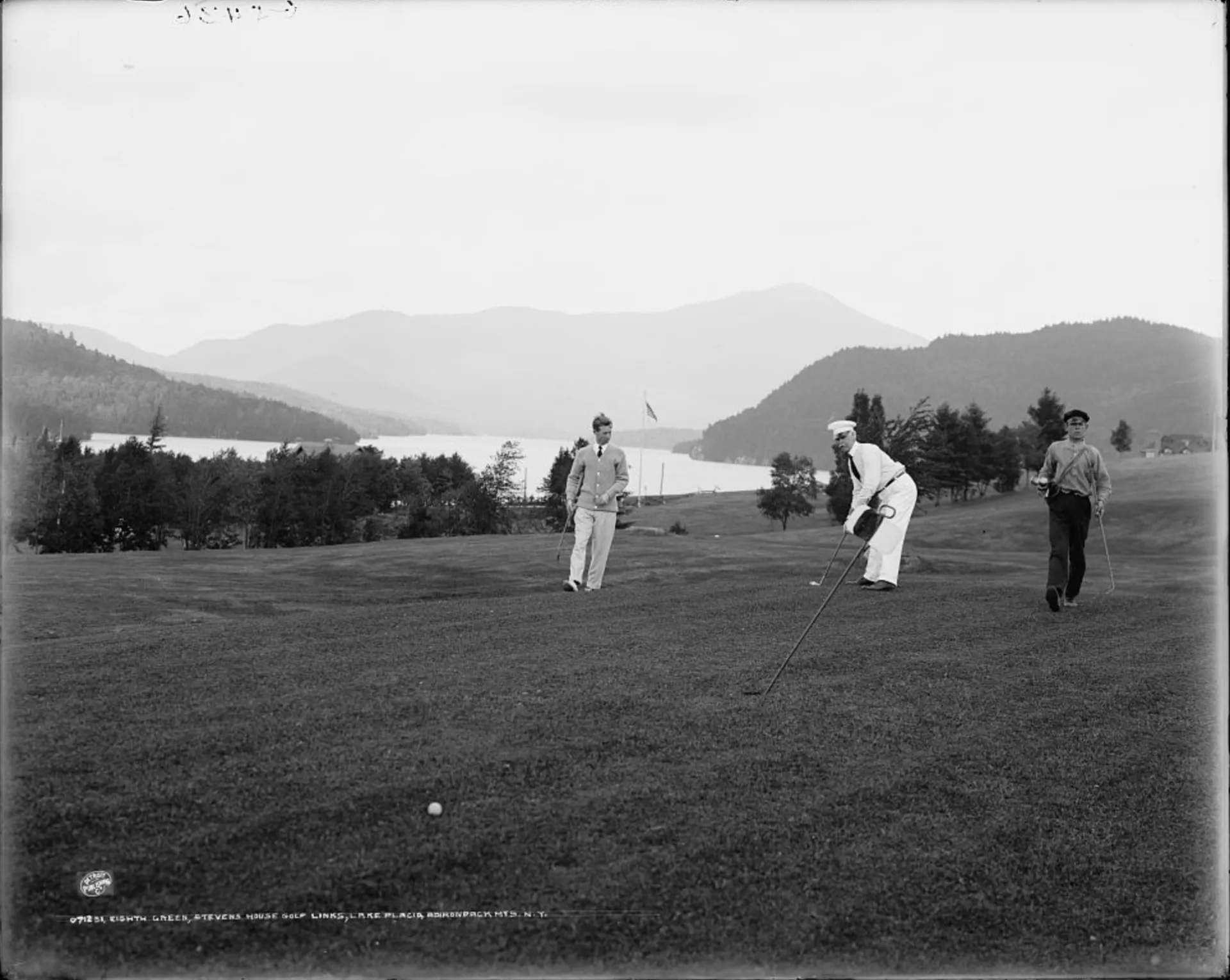 A vintage black and white image showing three men golfing with a lake and mountains in the background.